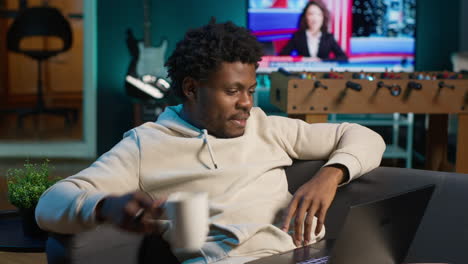 young adult working remotely from his loft apartment on a laptop. black freelancer focusing on tasks, blending productivity and relaxation while browsing the web from a cozy sofa. camera b.