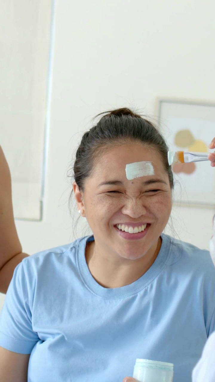 Vertical video: Getting woman in blue tee smiling, right hand applying mask for home spa with brush