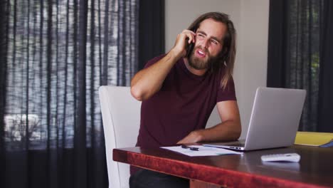 hombre caucásico hablando en un teléfono inteligente mientras usa una computadora portátil en casa