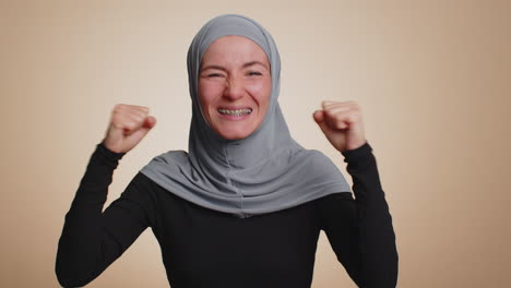 Stylish-woman-in-hijab-shouting,-raising-fists-in-gesture-I-did-it,-celebrating-success,-winning