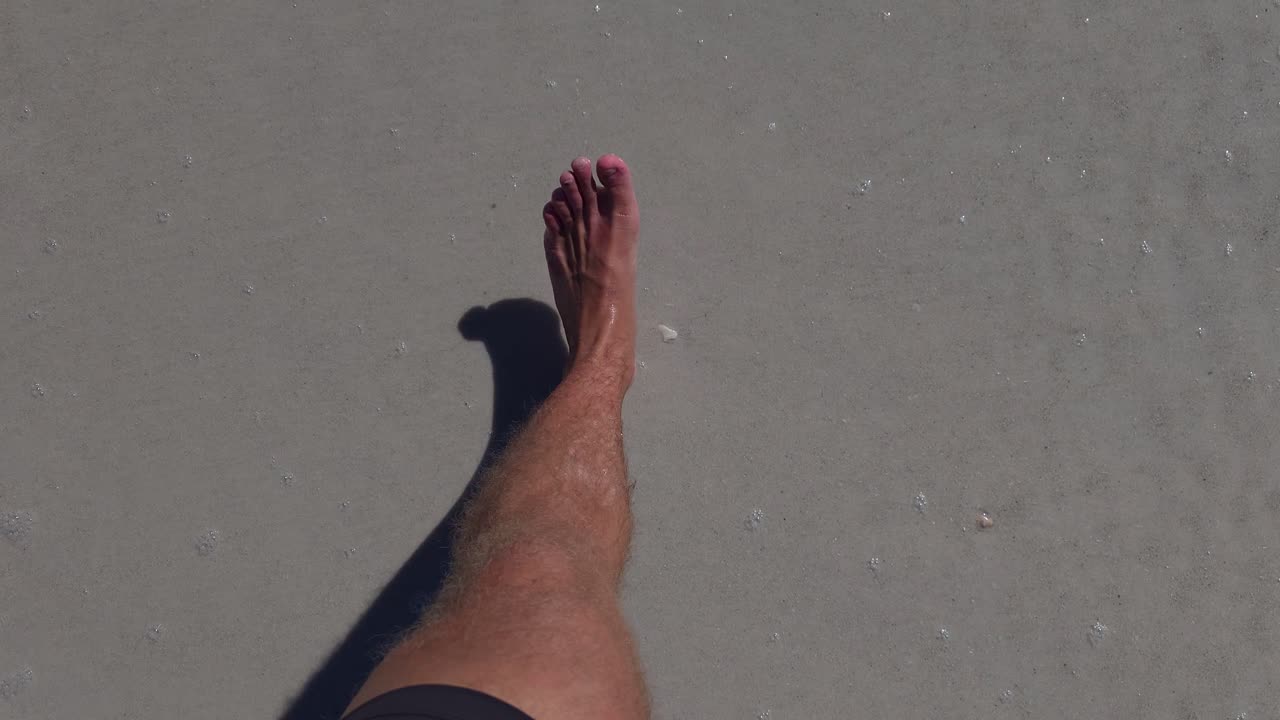 A barefoot person walks calmly along smooth wet sand near the shoreline, leaving gentle footprints while small bubbles of sea foam dot the surface.