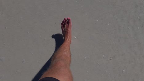 a barefoot person walks calmly along smooth wet sand near the shoreline, leaving gentle footprints while small bubbles of sea foam dot the surface.