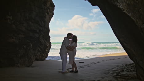 expectant father mother kissing standing sand beach in stone cliff shadow.