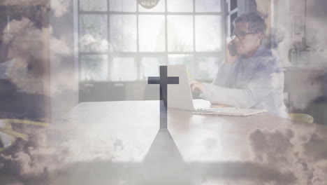 animation overlay of cross and clouds on person working on laptop at school desk