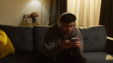 young man spending evening at home sitting on sofa wearing wireless headphones streaming music from mobile phone 3