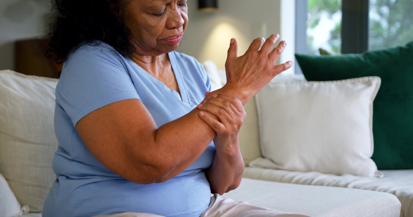 Anziana donna afroamericana che alza la mano destra, strofinandosi il polso destro per il dolore, sul divano con cuscini.