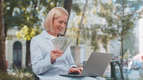 happy mature caucasian businesswoman ceo holding cash and using laptop computer on city street