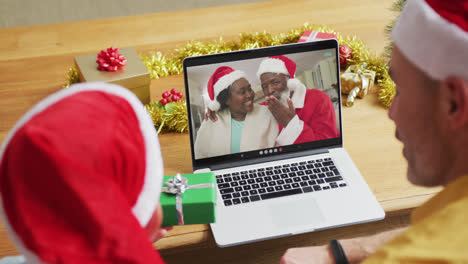 Padre-E-Hijo-Caucásicos-Con-Gorros-De-Papá-Noel-Usando-Una-Computadora-Portátil-Para-Una-Videollamada-Navideña-Con-Una-Pareja-En-La-Pantalla