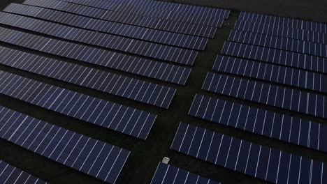 Sideways-aerial-view-of-solar-panel-station-with-long-rows-of-power-supply-reflective-absorbing-renewable-energy-at-sunset-with-the-low-sun-behind-electricity-towers-at-the-horizon