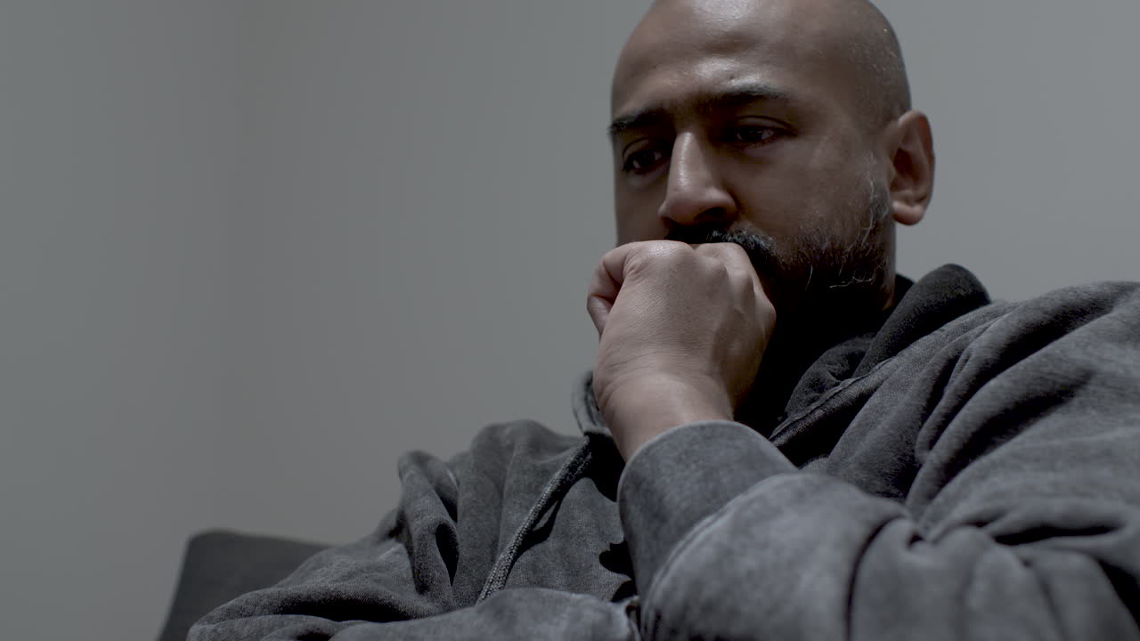 un primer plano de un hombre indio ansioso sumido en sus pensamientos con los ojos mirando al espacio, su mano apretada cubriendo su rostro mientras se mece hacia adelante y hacia atrás respirando profundamente