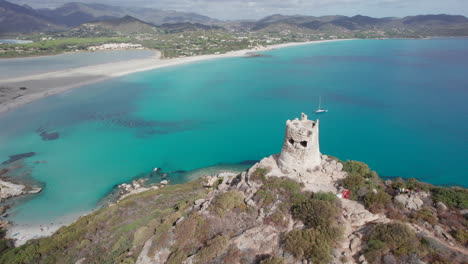 Orbital-aerial-view-of-the-Torre-di-Porto-Giunco-in-Cape-Carbonara-with-the-beautiful-beach-in-the-background-and-a-sunny-day