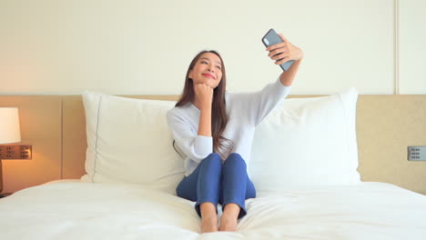 Una-Sonriente-Chica-Asiática-De-Aspecto-Agradable-Mirando-La-Cámara-De-Un-Smartphone-Moderno-Y-Tomándose-Selfies-Mientras-Se-Sienta-En-La-Cama-En-Una-Habitación-De-Hotel