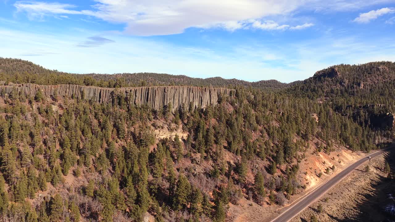 Et vidvinkel landskabsbillede af en bil, der kører på en afsidesliggende asfaltvej. Ruten snor sig forbi en monumental klippe af naturlige basaltsøjler og en frodig bjergskov.