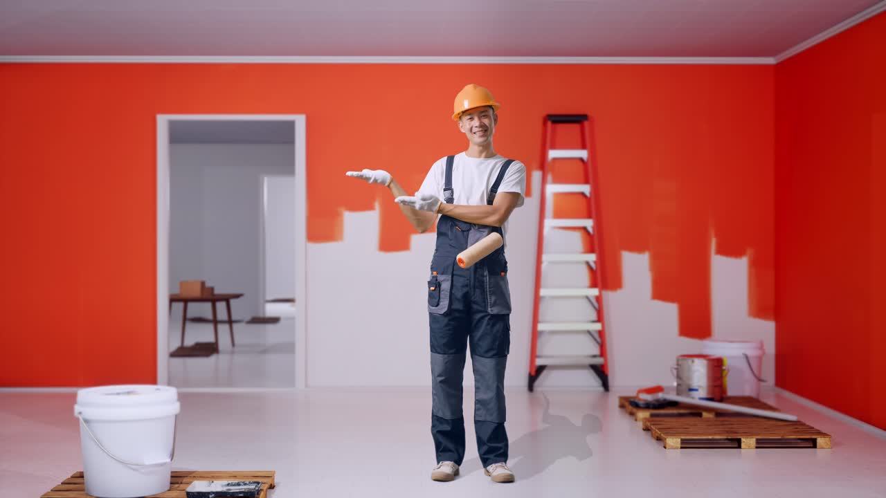 cuerpo lleno de pintor hombre asiático con casco de seguridad sonriendo y señalando a un lado mientras estaba de pie con la pared de pintura en la habitación