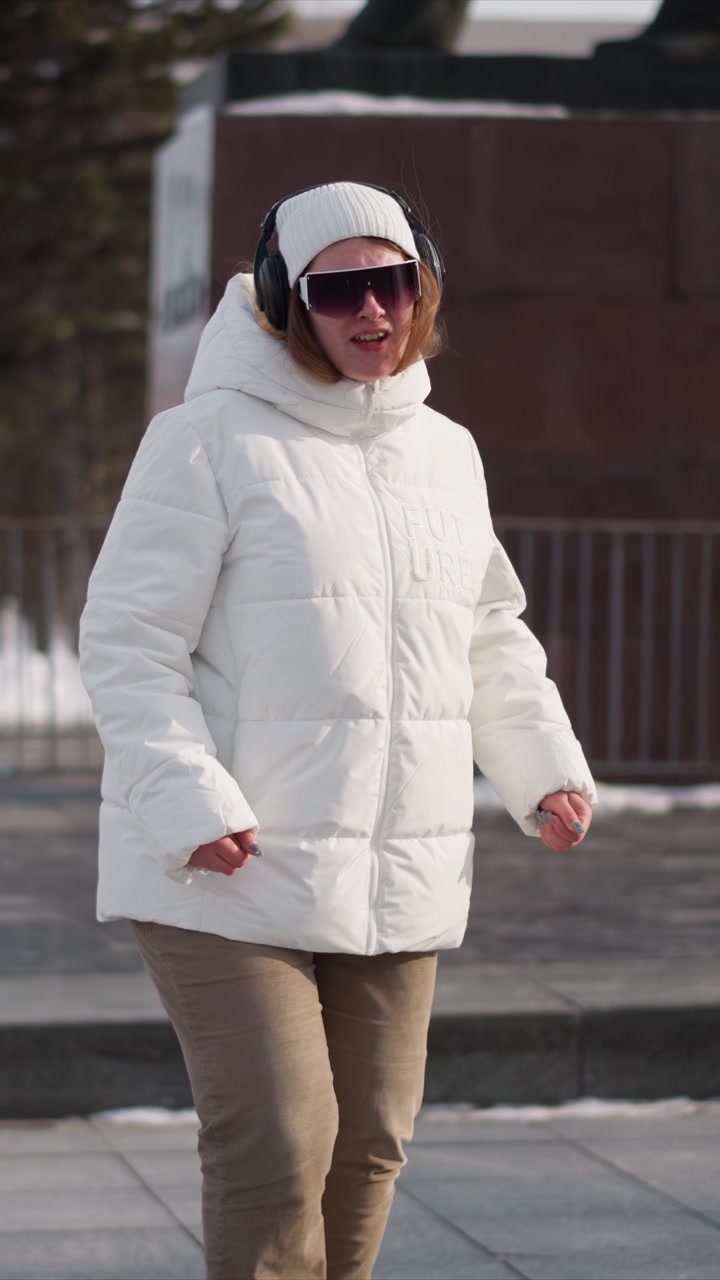 Woman wearing white puffer jacket beige pants and black headphones walks outdoors in urban setting with buildings visible. Woman gestures naturally as woman moves through environment