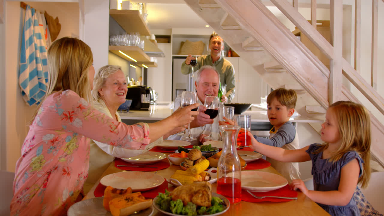 Kaukasisk familie på tværs af generationer, der skåler med drinksglas ved spisebordet derhjemme 4K
