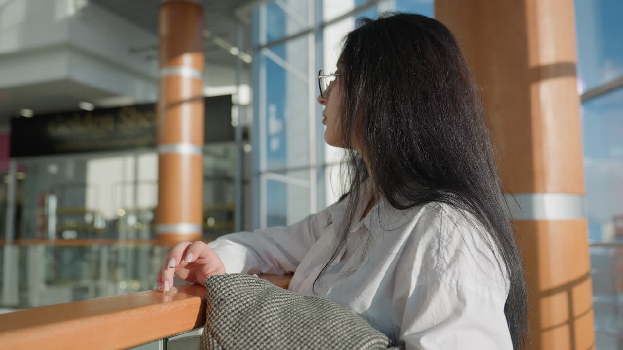 Side view of woman with long dark hair and glasses resting thoughtfully on wooden frame in bright shopping mall, holding jacket, gazing away, with elegant surroundings