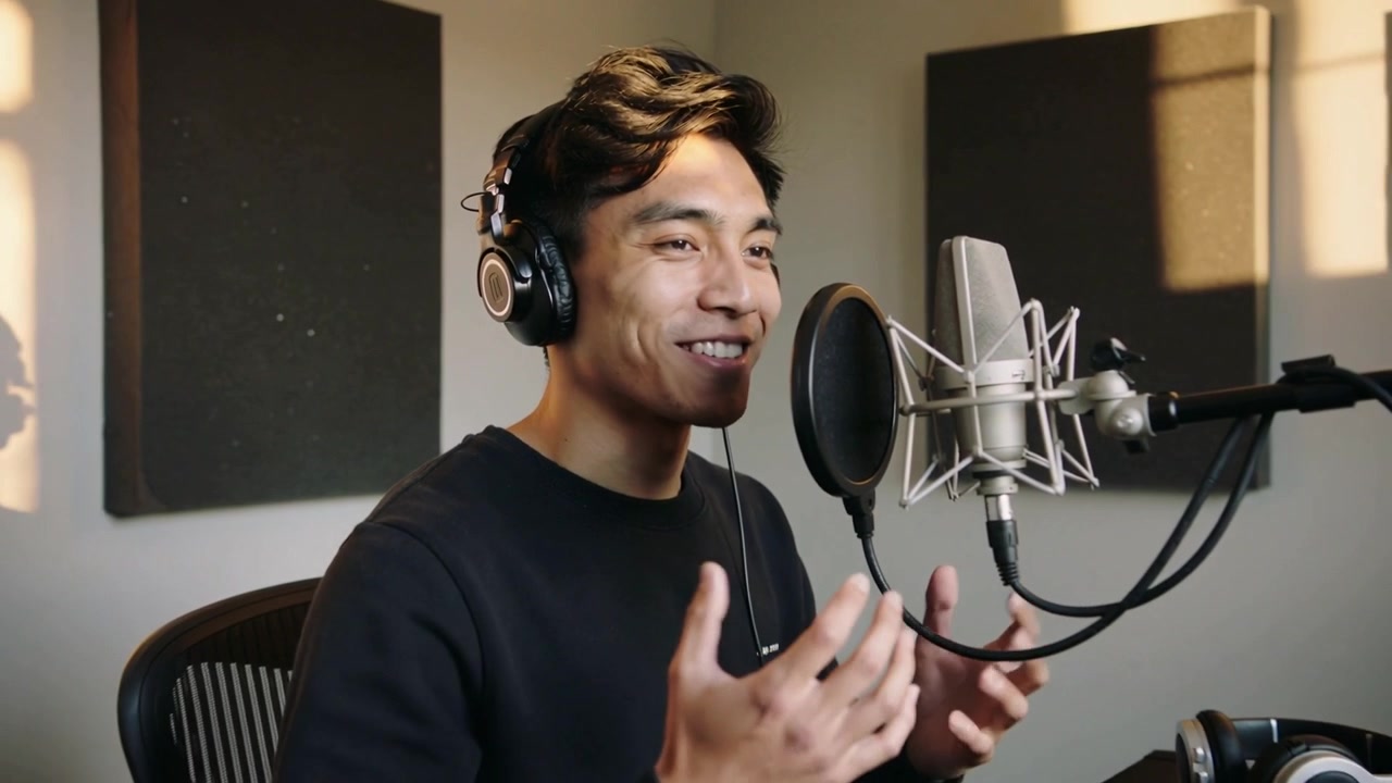 Cantante asiatico con maglietta scura che gesticola in studio mentre registra in un microfono, con la telecamera che si allontana.