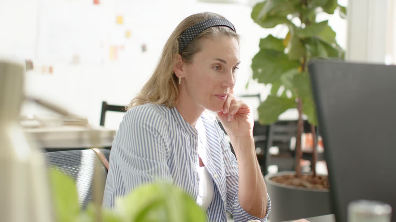 Mid-adult woman typing pausing to review screen, resting hand on chin, scanning keyboard at desk
