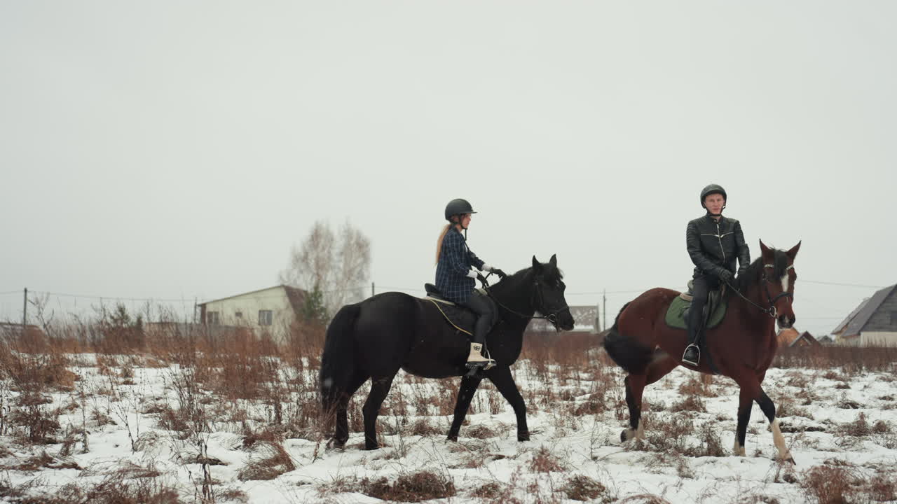 Due cavalieri bianchi in piedi su un campo innevato, l'istruttore che dà indicazioni verbali durante una pausa di formazione in tandem, l'allievo che ascolta accanto al cavallo, con i caschi indossati, le selle strette, case del villaggio in lontananza, tranquillo.