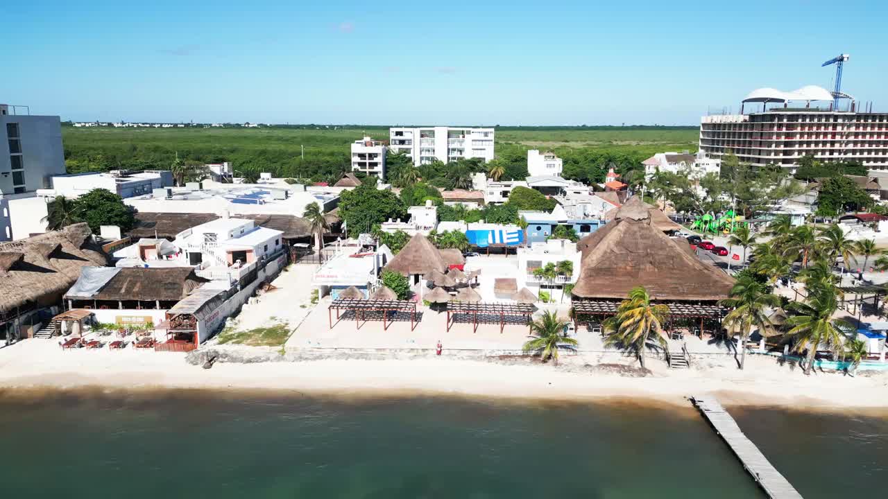 Aerial pullback revealing the sandy beachfront, palm-lined shoreline, and pier in Puerto Morelos, Mexico, with coastal buildings and clear Caribbean waters coming into view