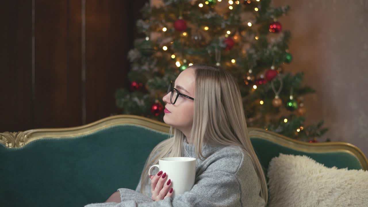 A young woman wearing glasses and a gray sweater enjoys a warm drink while sitting on a vintage green sofa near a beautifully decorated Christmas tree