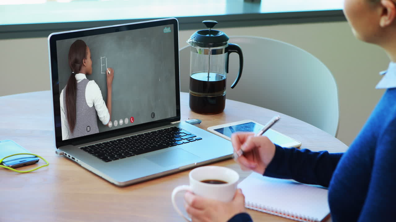 Caucasian female student using laptop on video call with female teacher, making notes
