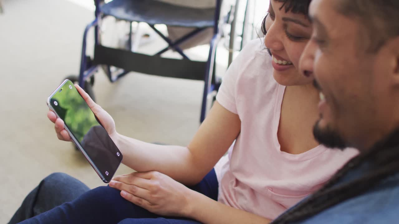 feliz pareja biracial en el sofá usando un teléfono inteligente con espacio de copia, su silla de ruedas en el fondo