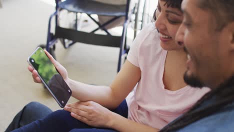 feliz pareja biracial en el sofá usando un teléfono inteligente con espacio de copia, su silla de ruedas en el fondo