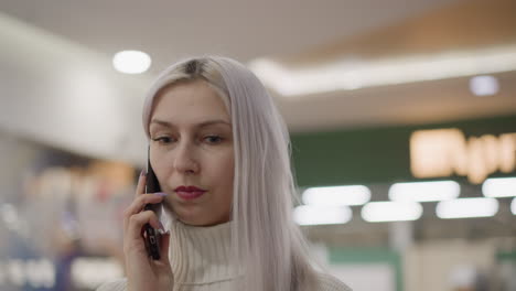 close up of trendsetting woman with pastel hair holding smartphone to ear listens thoughtfully in bustling shopping mall environment blurred shoppers and ceiling lights form dynamic retail atmosphere