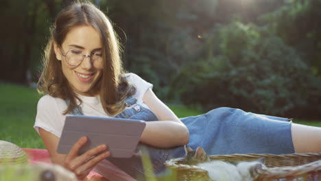 young caucasian woman in glasses lying on a blanket in the park tapping and scrolling on a tablet