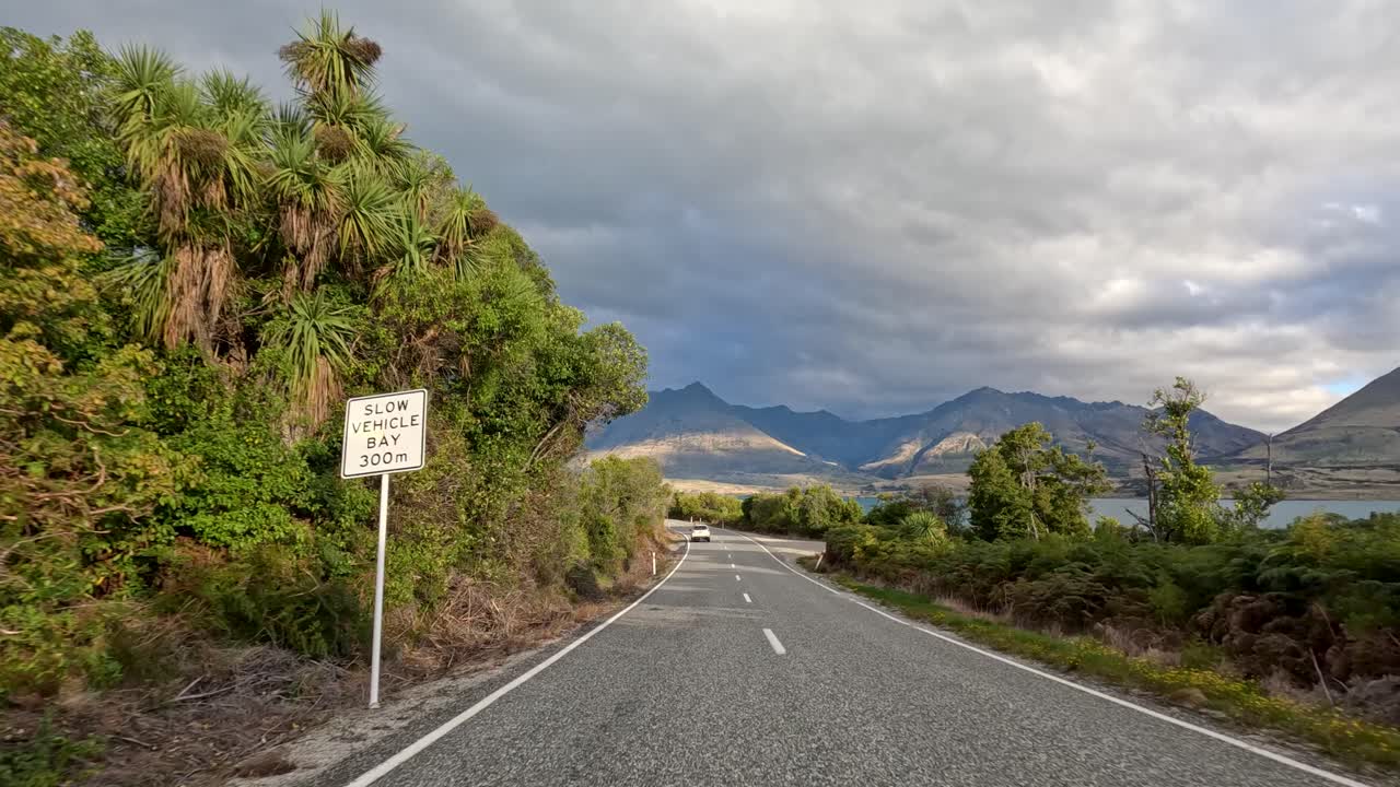 Forward-facing car camera captures winding road, lush greenery, and dramatic mountains under cloudy daylight