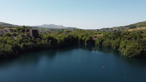 Aerial-rising-tilt-view-Welsh-woodland-valley-slate-mining-shaft-and-quarry-lake-revealing-Snowdonia-mountains