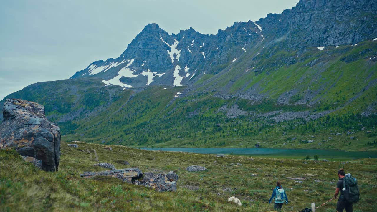 SørTverrfjord, Loppa, Finnmark, Norja – Kaksi retkeilijää kävelee koiriensa kanssa rosoisten, lumiraidallisten huippujen alla tyynen vuoristojärven rannalla – Staattinen kuva.