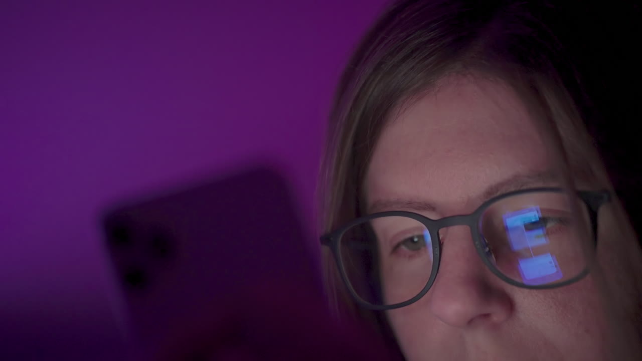 Reflection of phone screen on girl's glasses as she browses social media on smart phone late at night