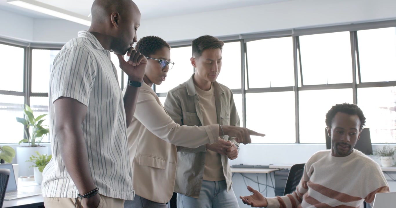 Diverse coworkers leaning around seated African man at desk over tablet content, clarifying task