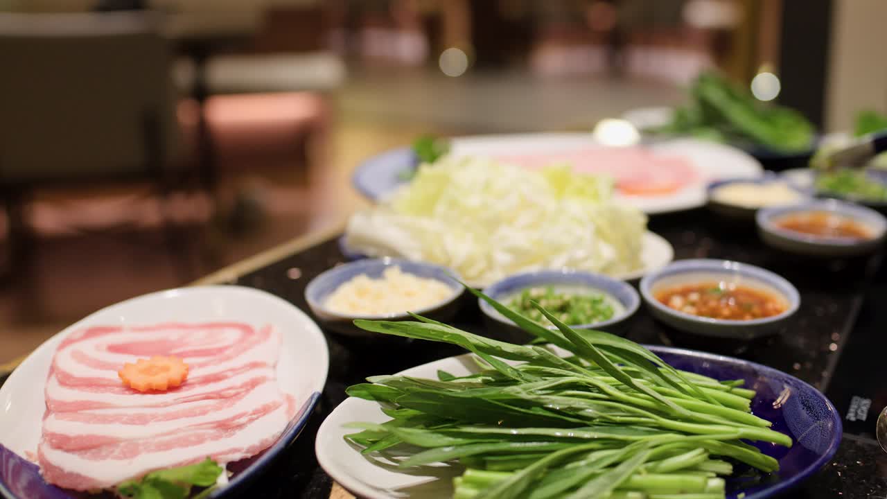 Hand arranges vegetables and sliced meat for hotpot on table under warm indoor lighting