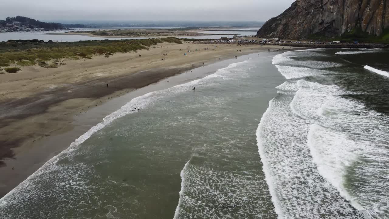 Aerial shot of waves crashing onto the sand at Morro Beach in California on a cloudy day in California. The beach sits in the foreground while the bay, Morro Rock, and sand dunes in the background.