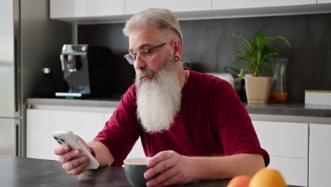 Un-Hombre-Serio,-De-Pelo-Gris-Y-Barba-Frondosa,-De-Edad-Avanzada,-Con-Gafas-Y-Camisa-Roja,-Se-Sienta-A-Mirar-Una-Transmisión-En-Un-Teléfono-Inteligente-De-Una-Red-Social-Durante-Su-Desayuno-Y-Sonríe-En-Una-Cocina-Moderna.