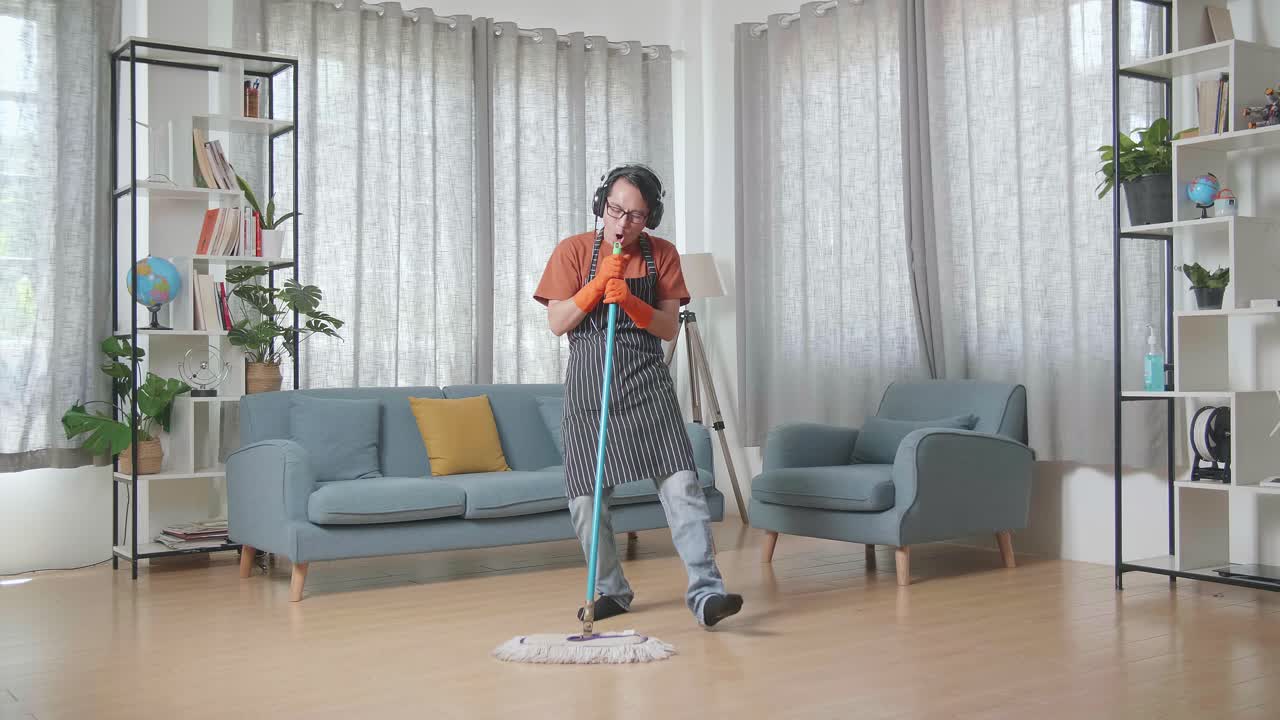 ama de casa asiática con un delantal usando auriculares, escuchando música y bailando mientras limpia el suelo en casa
