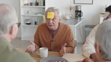 waist-up over shoulder of cheerful older people sitting at table, playing post-it note game, caucasian man asking questions of his character, answering, african woman smiling, applauding