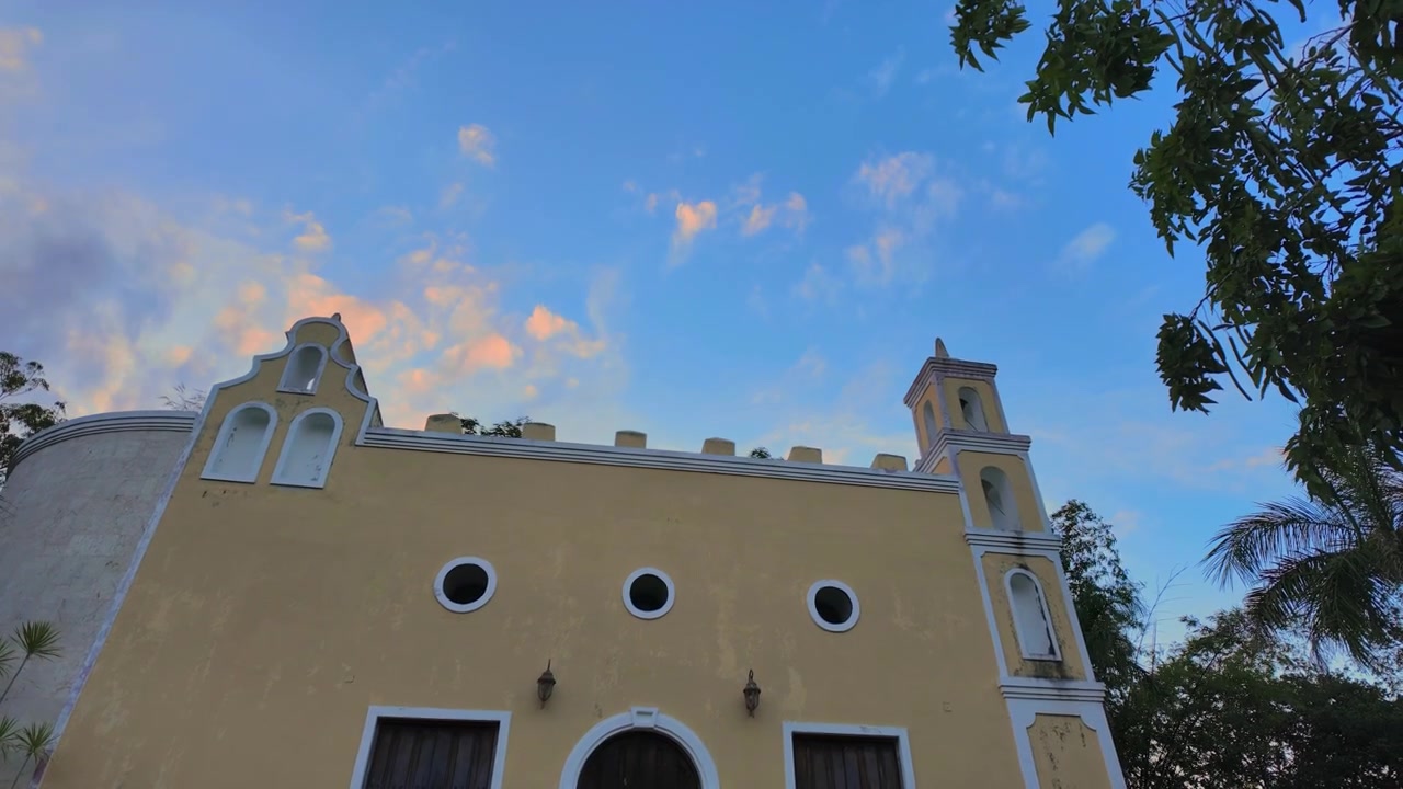 Yucatan, güney Meksika'da, çiftlik işçileri için inşa edilmiş kolonyal bir hacienda şapelinin soluk sarı cephesi ve küçük çan kulesi, pembe bulutlarla kaplı derin mavi bir akşam gökyüzüne karşı duruyor.