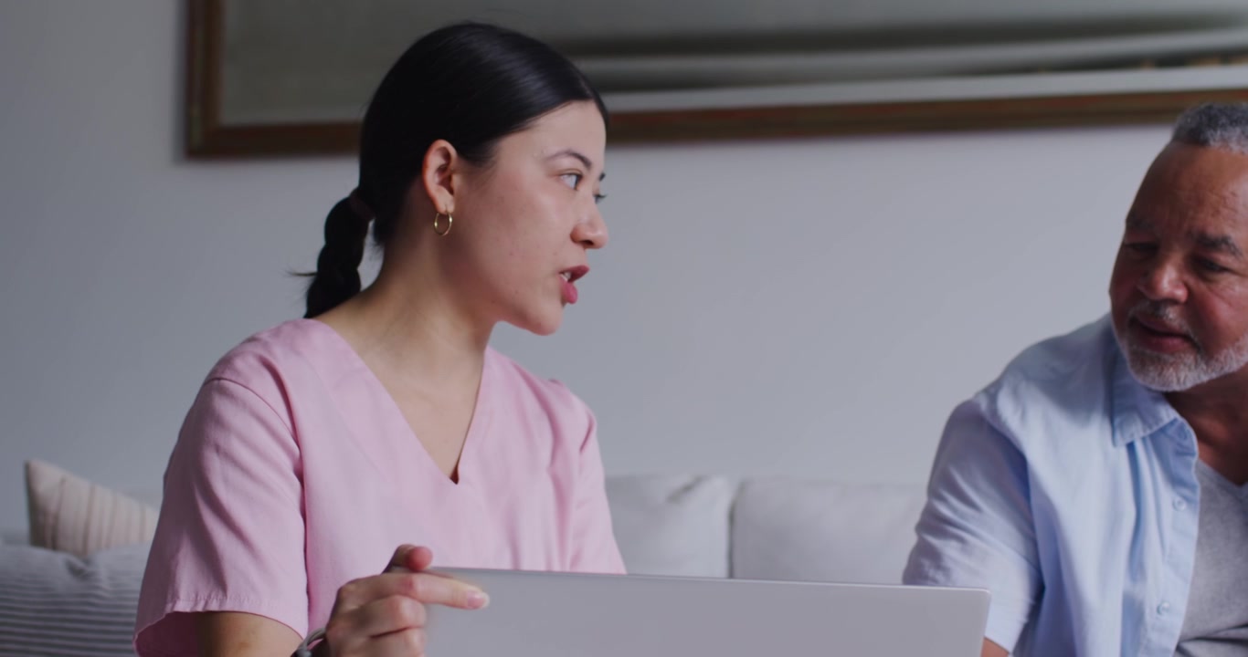 Diverse caregiver and senior man, woman in pink scrubs opening laptop and explaining care on sofa