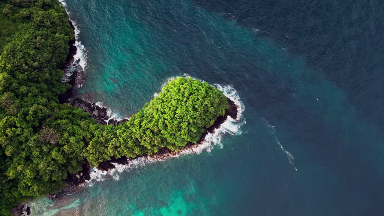 Drone footage of a lush green peninsula in Karangasem, Bali, surrounded by turquoise water and rocky shores. Waves crash beautifully along the coastline, showcasing Indonesia untouched coastal nature