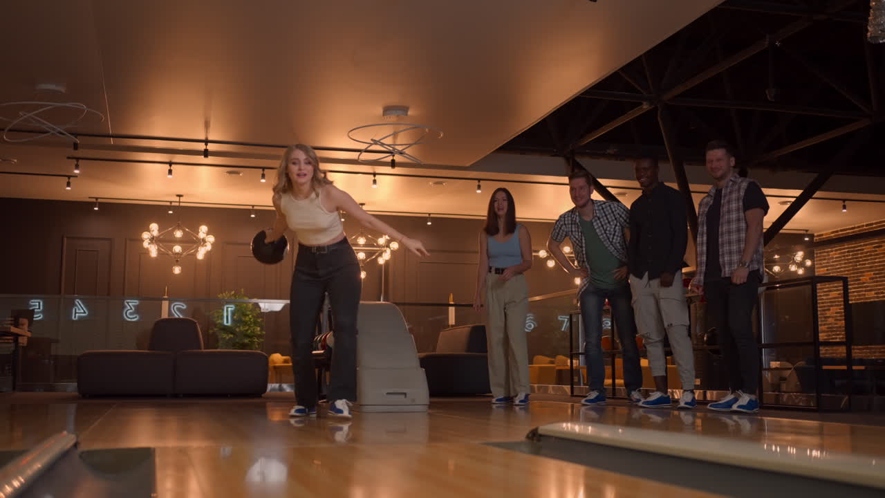A woman in a bowling alley throws a ball on the track and knocks out a shot in slow motion and jumps and dances joy. Friends support and clap with a smile. multi-ethnic friends play bowling together.