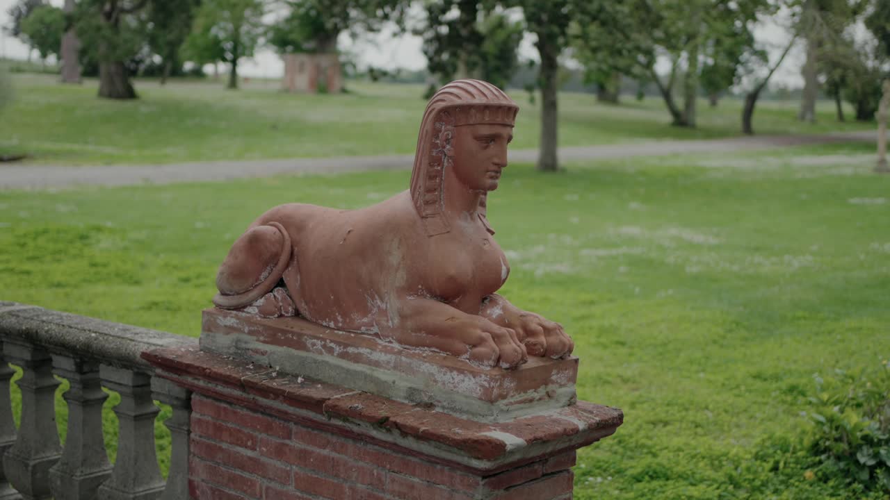 A terracotta sphinx statue on a brick pedestal set in a green park, with trees and a path in the background