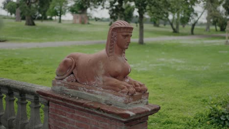 A-terracotta-sphinx-statue-on-a-brick-pedestal-set-in-a-green-park,-with-trees-and-a-path-in-the-background