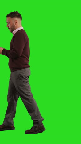 vertical video full length studio shot of businessman using mobile phone to message or browse online walking across green screen