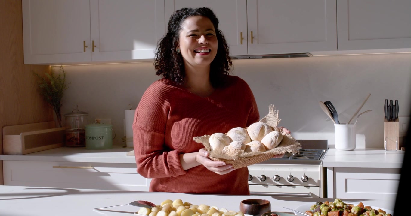 Mulher afro-americana com suéter cor ferrugem apresentando cesta de pães na ilha da cozinha, câmera gravando