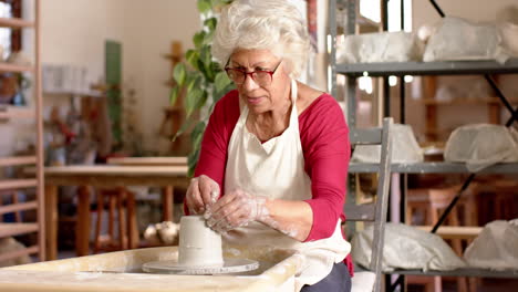 senior biracial female potter with gray hair using potter's wheel in pottery studio, slow motion